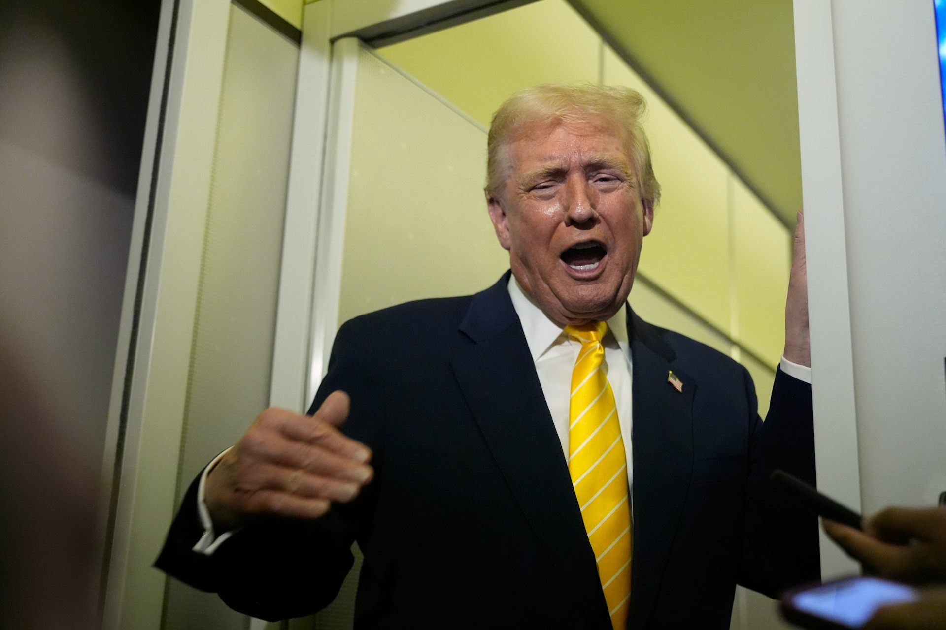 A man with orange-ish skin tone and fluffy yellow-orange hair in a dark suit with a yellow tie gestures and grimaces as he speaks.