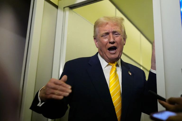 A man with orange-ish skin tone and fluffy yellow-orange hair in a dark suit with a yellow tie gestures and grimaces as he speaks.