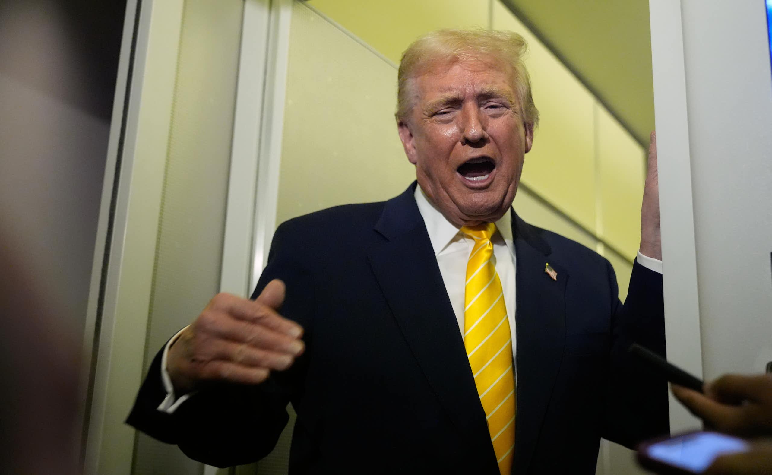 A man with orange-ish skin tone and fluffy yellow-orange hair in a dark suit with a yellow tie gestures and grimaces as he speaks.