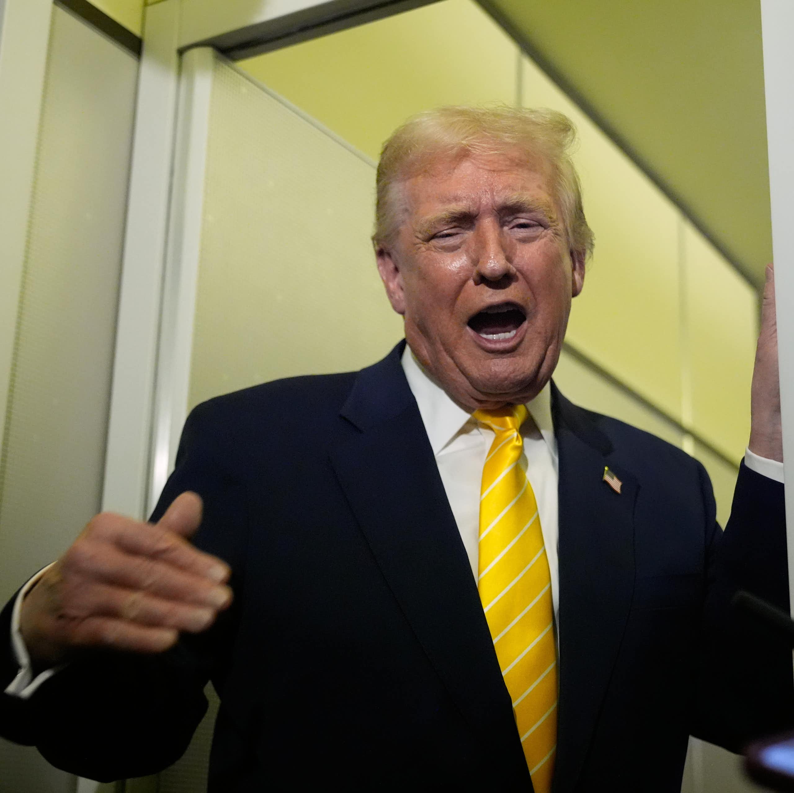 A man with orange-ish skin tone and fluffy yellow-orange hair in a dark suit with a yellow tie gestures and grimaces as he speaks.