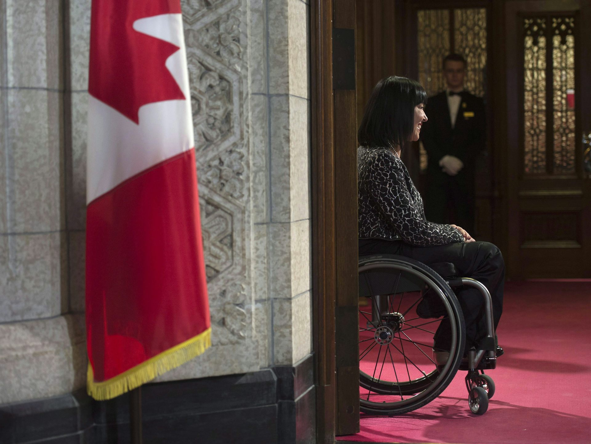 A dark-haired woman sitting in a wheelchair smiles in Parliament. A Canadian flag is in the foreground.
