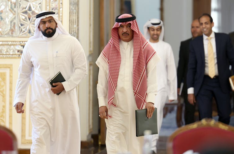 Three men in full Arab robes walk alongside two men in westernm-style suits.