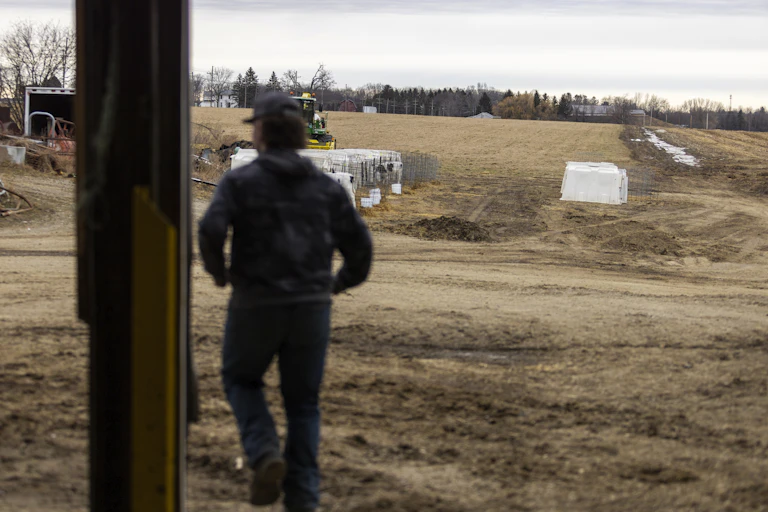 A person is seen from behind overlooking a large brown grassy area, trees and some farm machinery.
