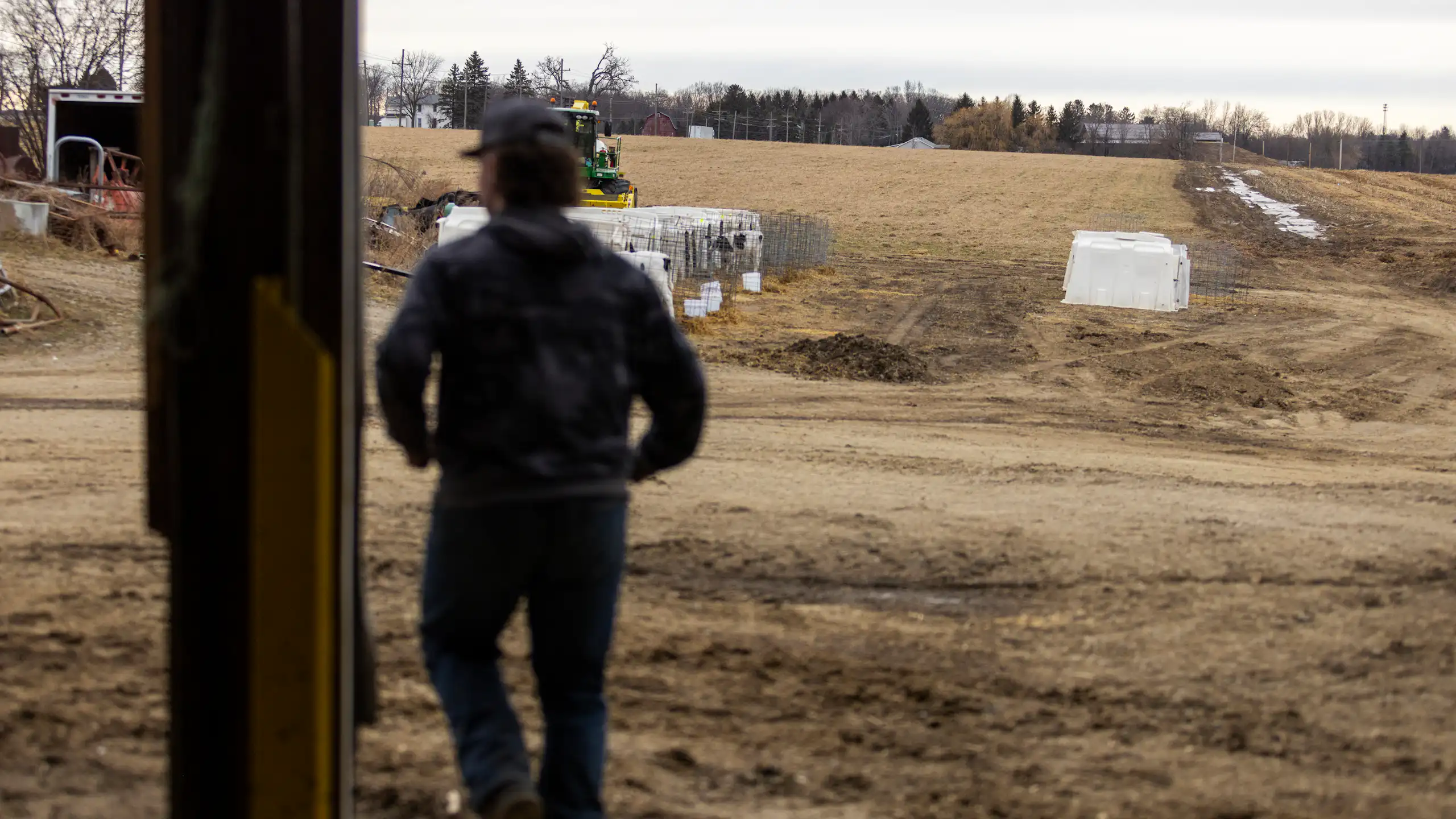 A person is seen from behind overlooking a large brown grassy area, trees and some farm machinery.
