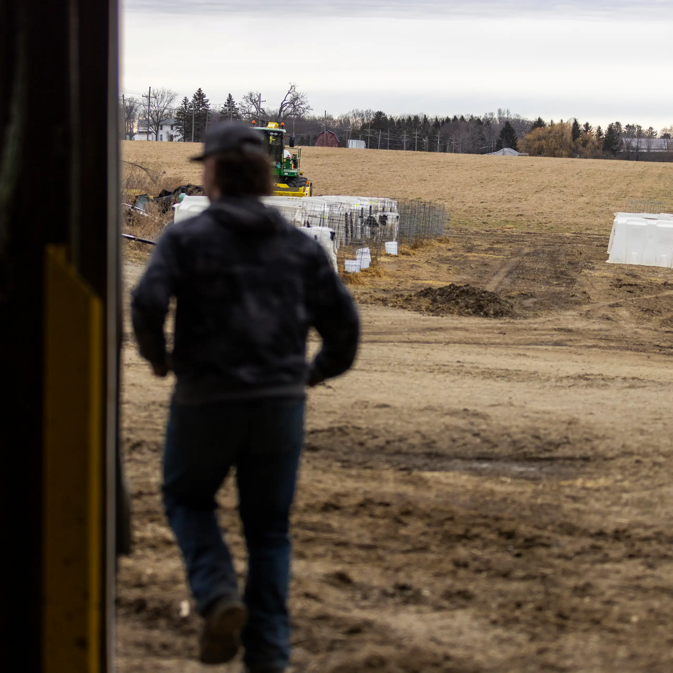 A person is seen from behind overlooking a large brown grassy area, trees and some farm machinery.