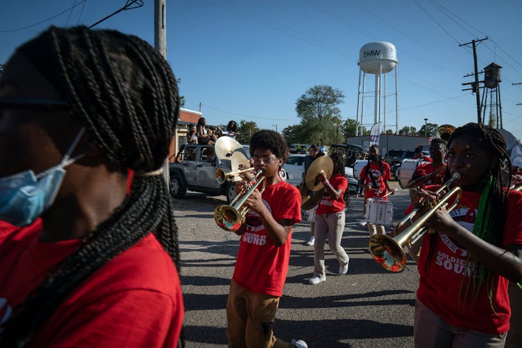 A group of young people wear red shirts and hold musical instruments, including trumpets, as they walk in the street on a blue-sky day.