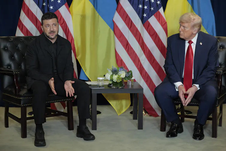 Two men sit on chairs in front of a number of flags.