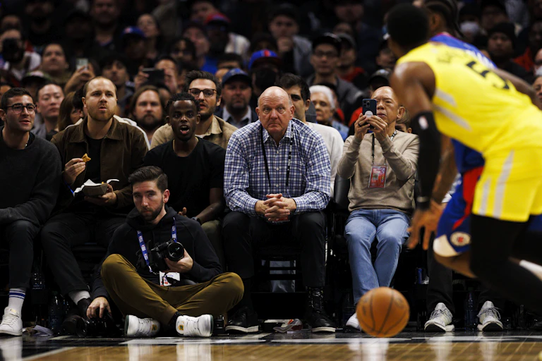 A man in the front row at a basketball game looks concerned watching two players battle for the ball.