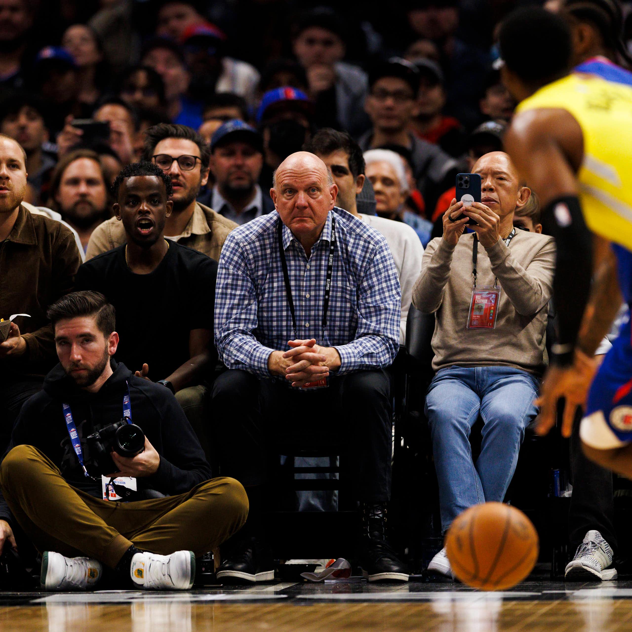 A man in the front row at a basketball game looks concerned watching two players battle for the ball.