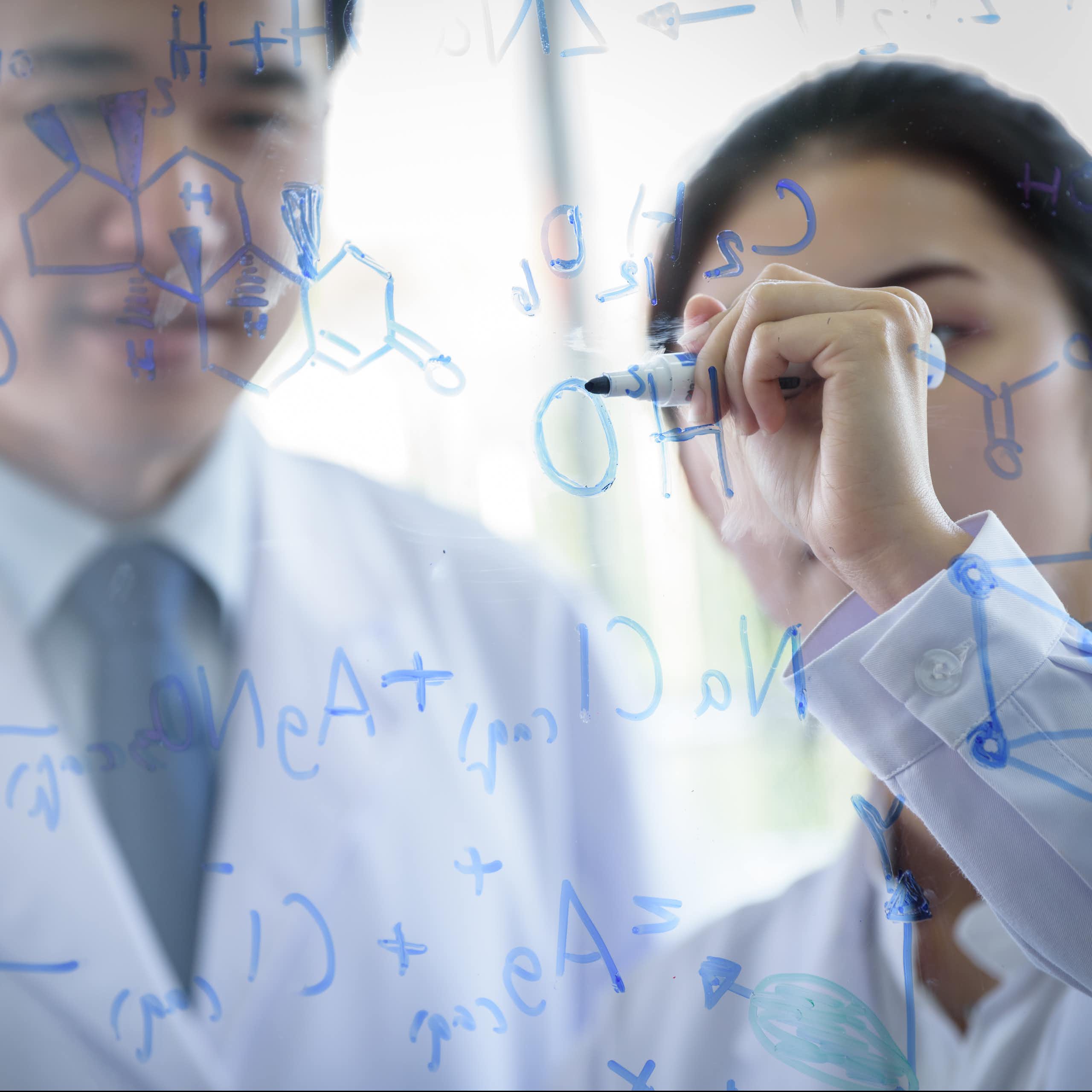 man and woman in white coats write chemical formulas with marker on clear glass
