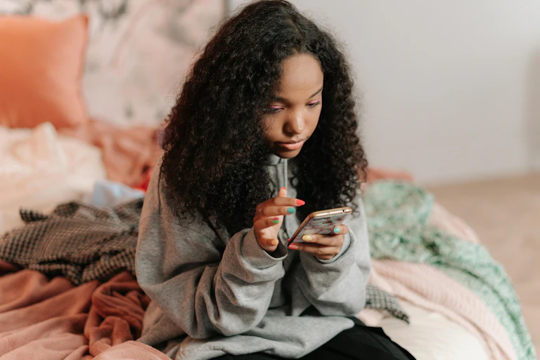 A teenager sits on a bed looking at her phone.