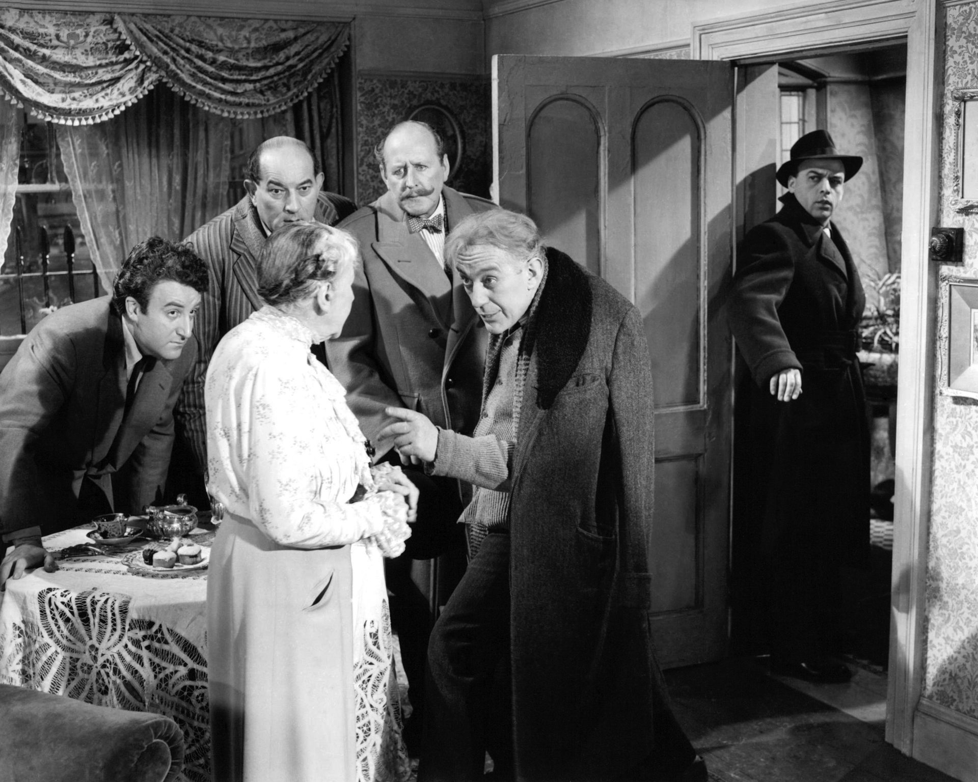 Black-and-white scene from film The Ladykillers showing an older woman surrounded by five men who watch her closely, with one man standing in a doorway in the background.