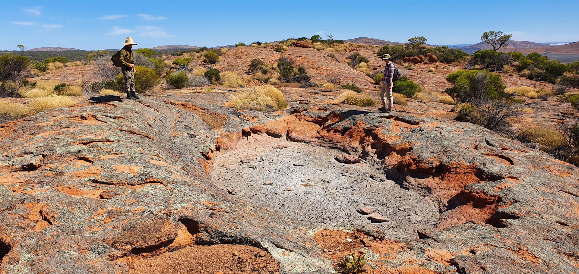 A dry rock-hole in a large granite outcrop. Two field workers stand nearby.