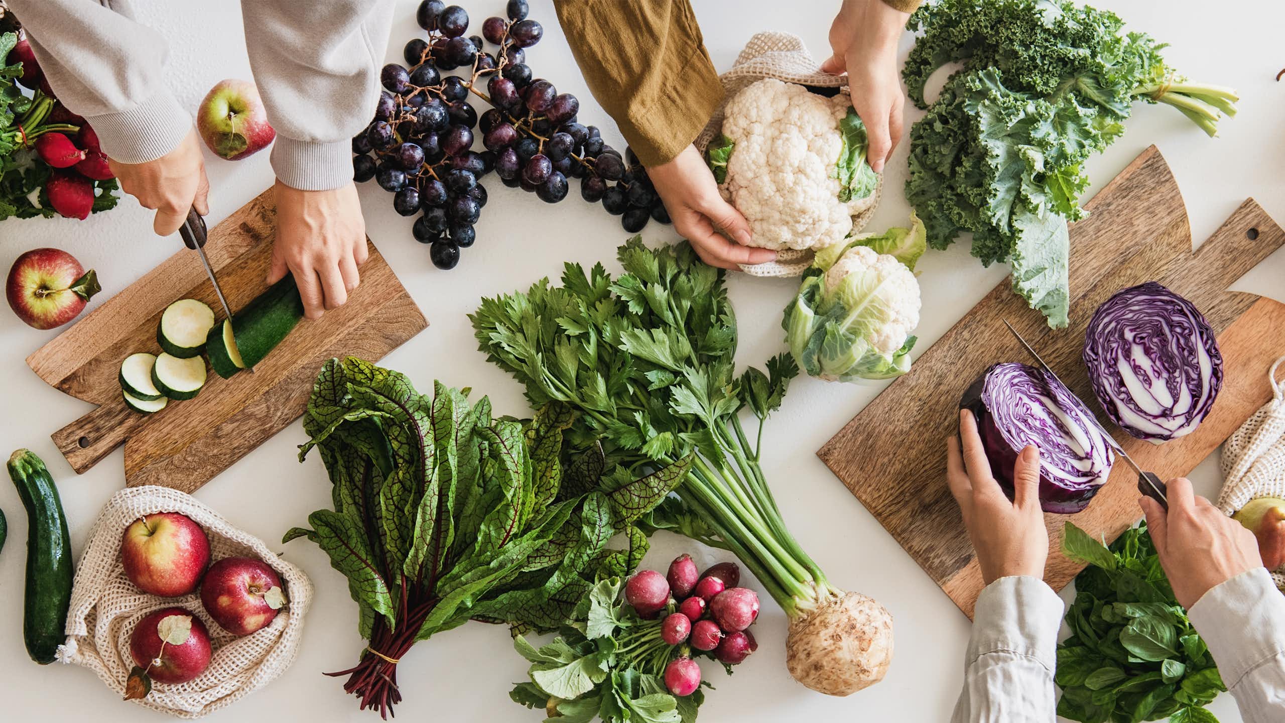 Des personnes sont en train de coupe des légumes (choux, courgettes, etc.) sur une table remplie de fruits et légumes verts.