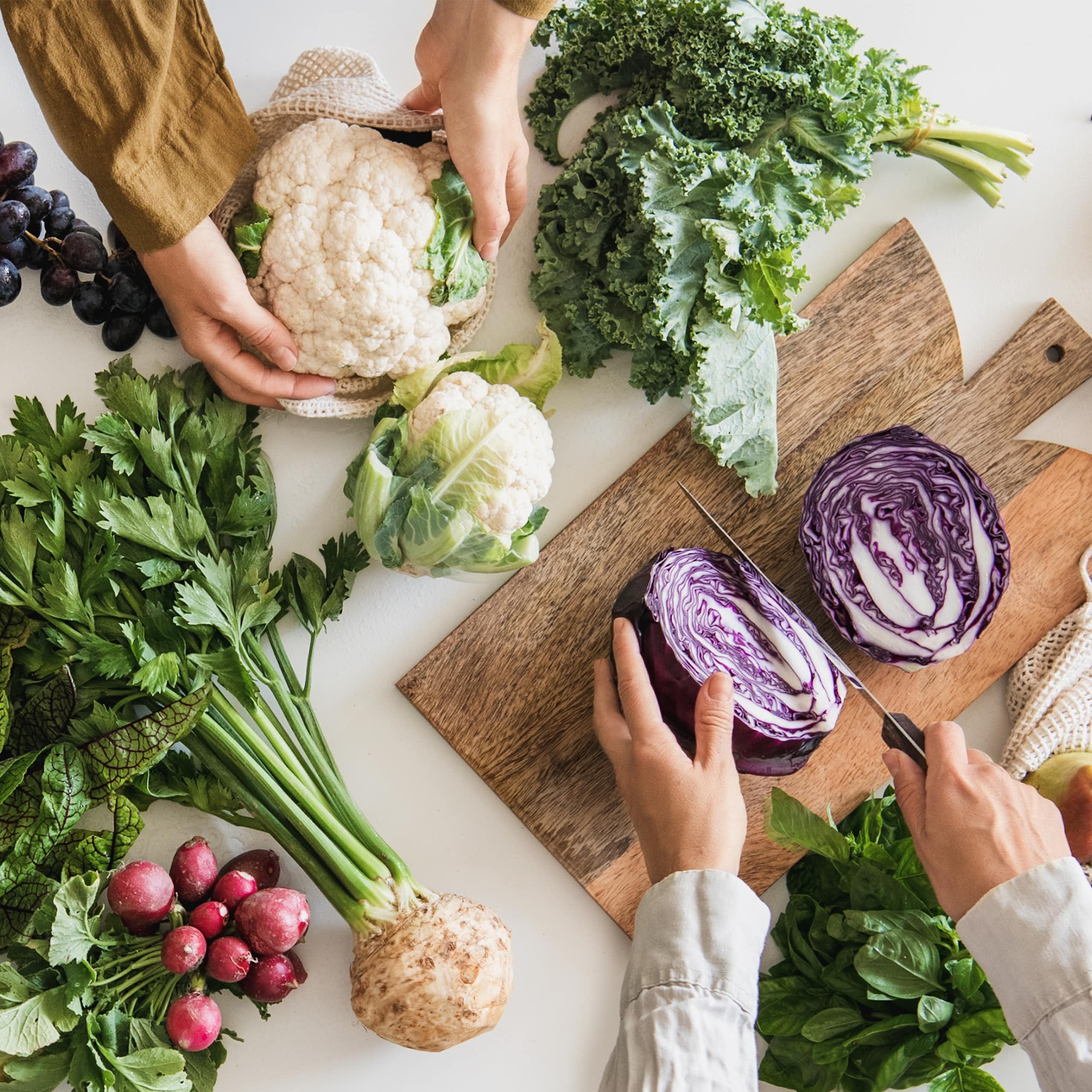 Des personnes sont en train de coupe des légumes (choux, courgettes, etc.) sur une table remplie de fruits et légumes verts.