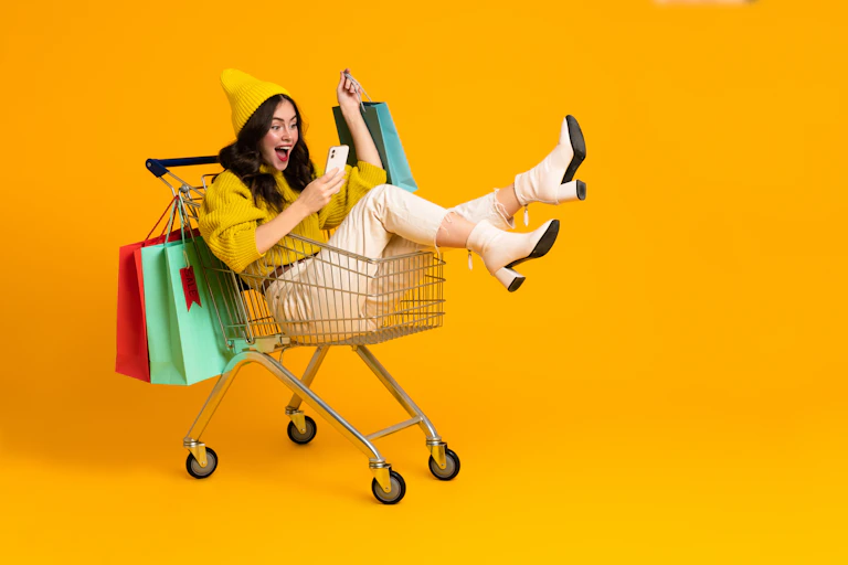 Yellow background, woman wearing yellow hat and jumper sitting in shopping trolley with shopping bags, looking at her phone.