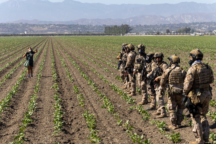A person standing in a field raises her hands as a line of people dressed as soldiers approaches.