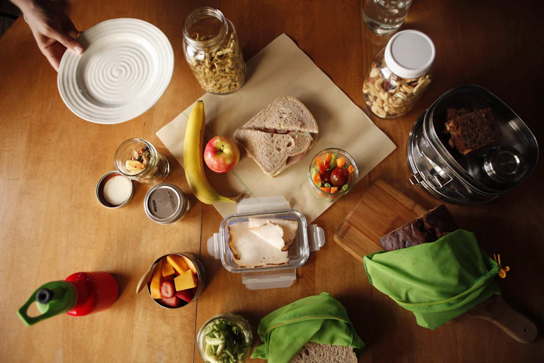 A sandwich sitting on a wax wrap on a table beside different foods and glass containers.