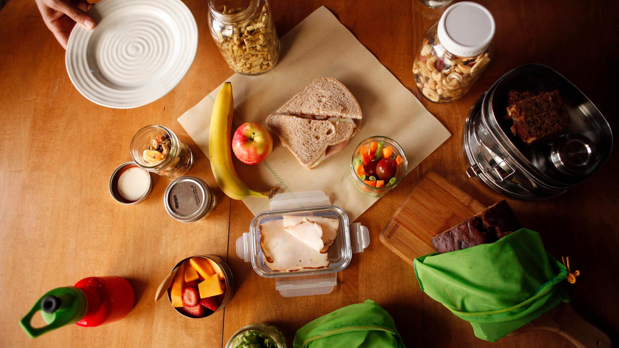 A sandwich sitting on a wax wrap on a table beside different foods and glass containers.
