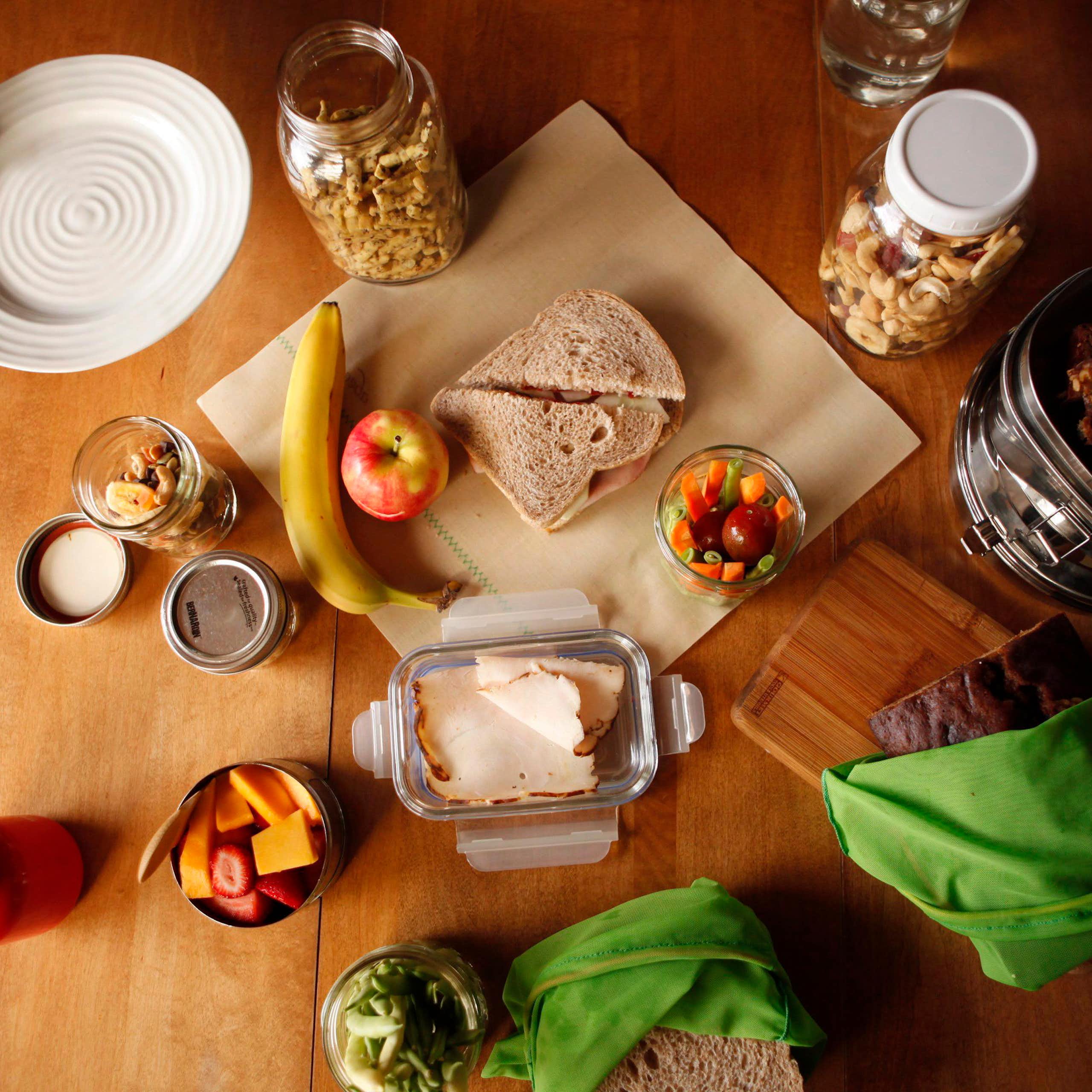 A sandwich sitting on a wax wrap on a table beside different foods and glass containers.