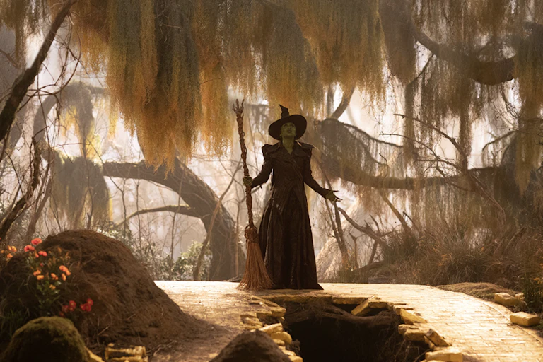 A witch in a black dress and pointy hat in a sinister looking wood with her broomstick.