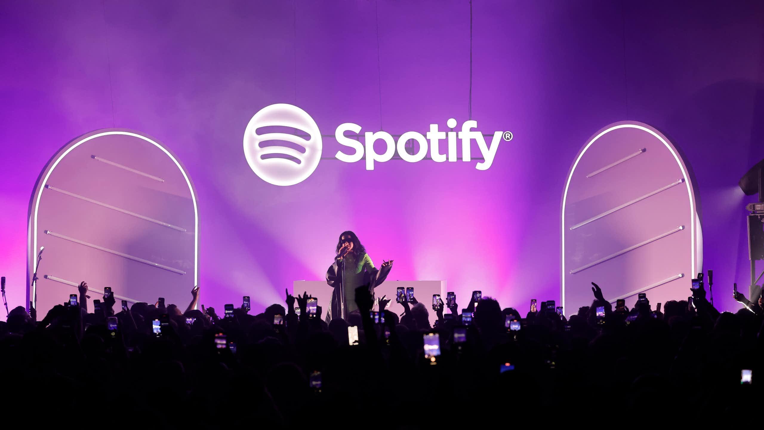 A young woman performs on a stage with a purple backdrop behind her in front a large crowd.