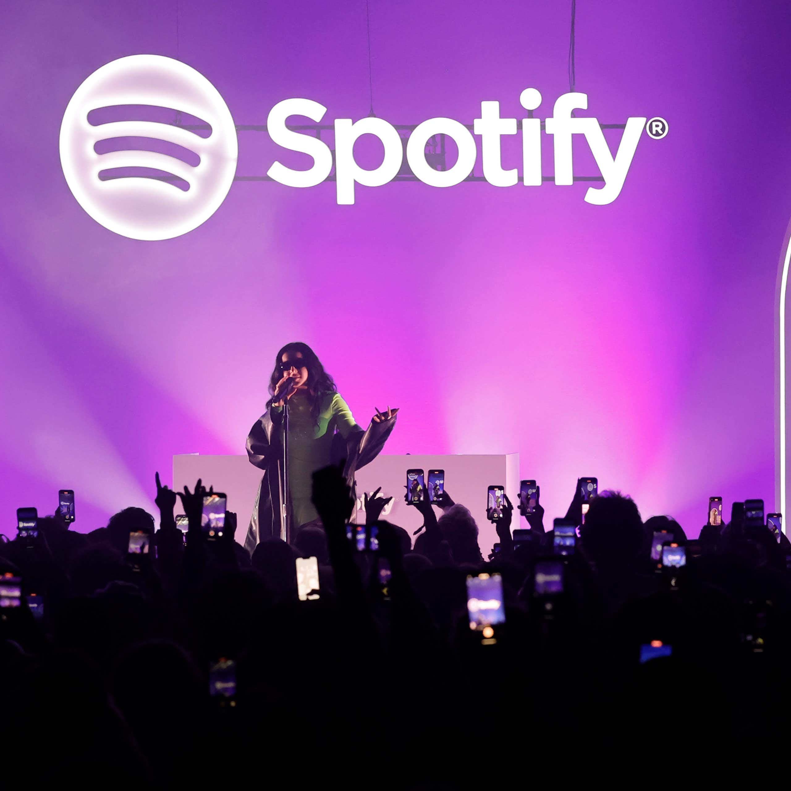 A young woman performs on a stage with a purple backdrop behind her in front a large crowd.