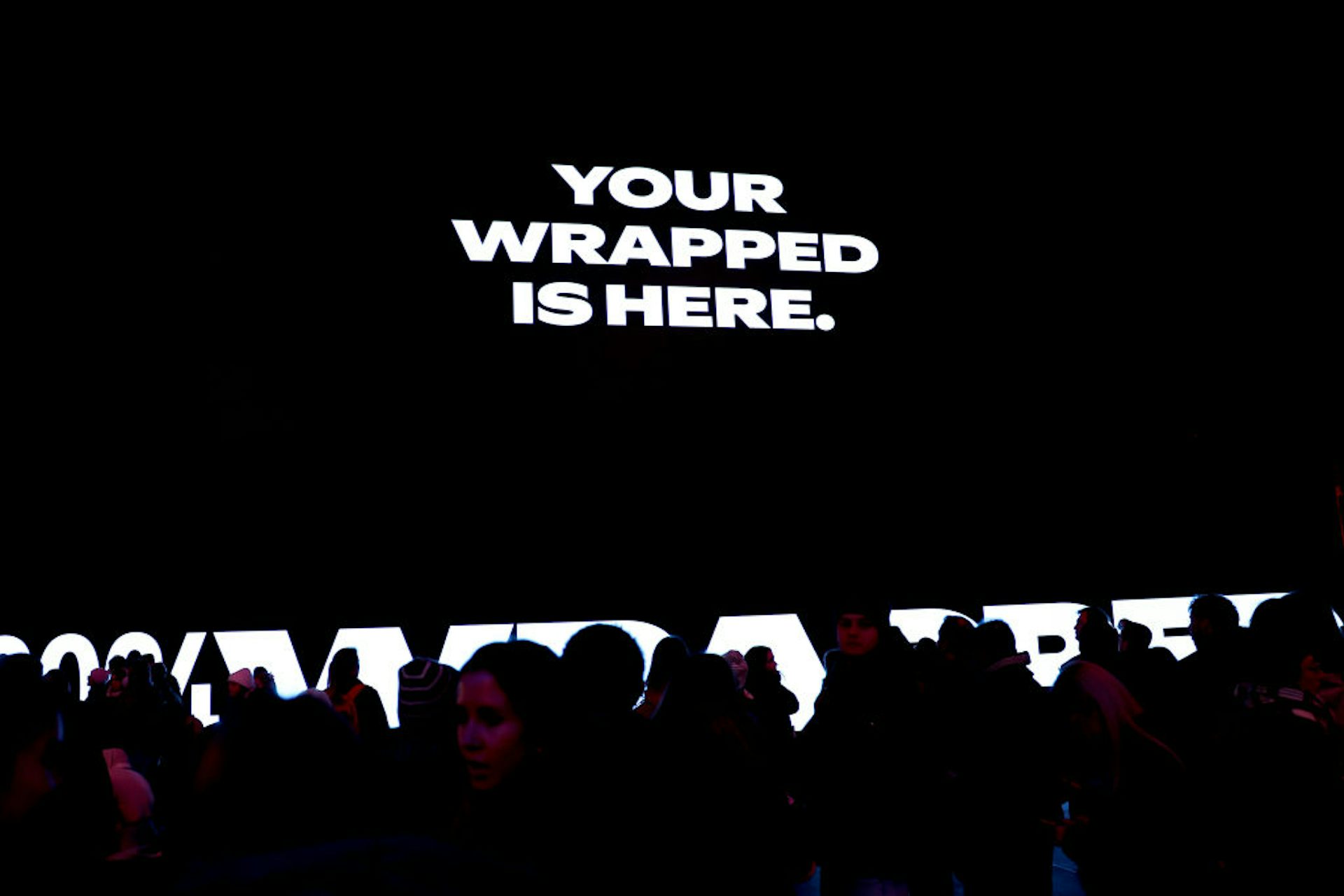 Text reading 'YOUR WRAPPED IS HERE.' is projected onto a black backdrop, visible over a silhouette of a large crowd.