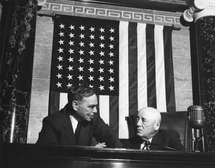 Two men at a desk, speaking, with an American flag hung behind them.