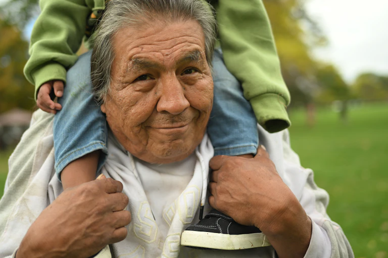 An older man with greying hair smiles as he holds a child on his shoulders.