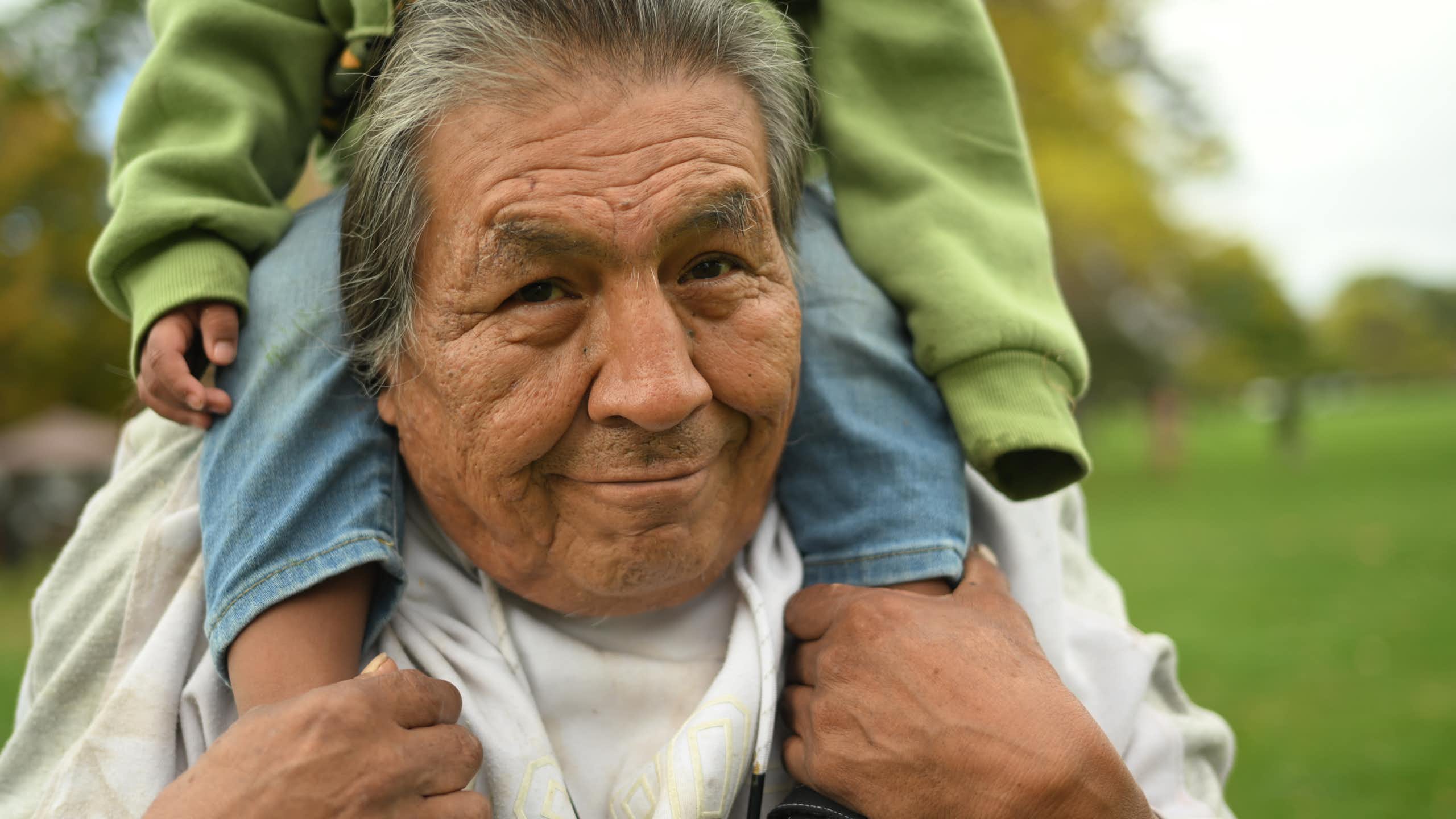 An older man with greying hair smiles as he holds a child on his shoulders.