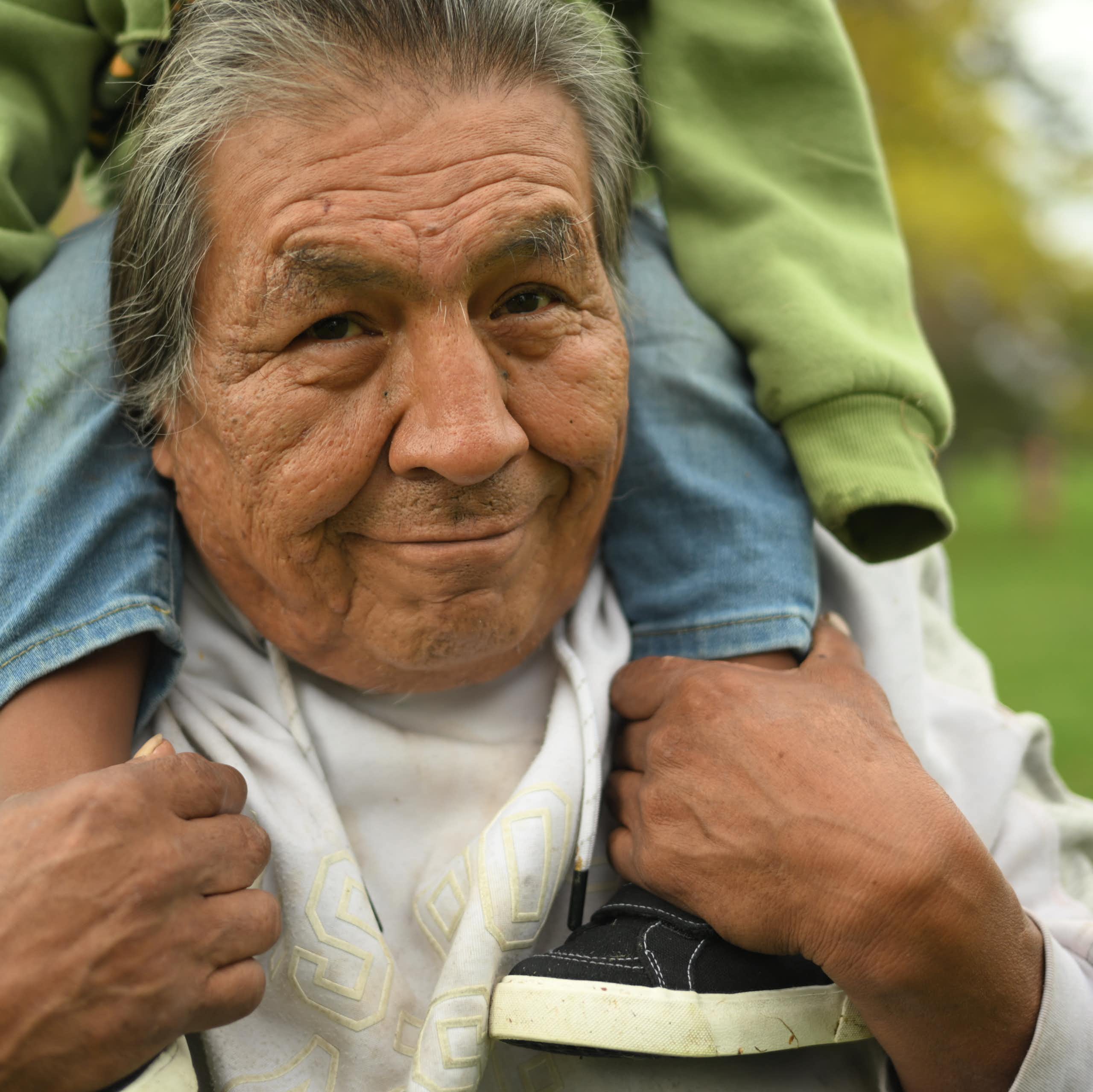 An older man with greying hair smiles as he holds a child on his shoulders.
