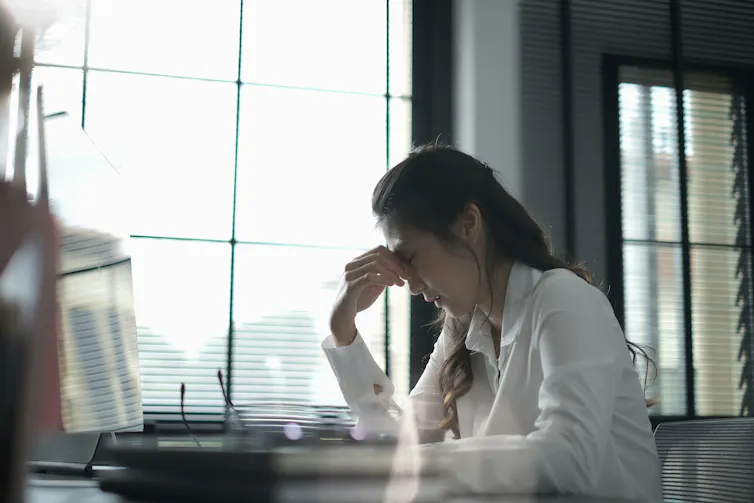 A woman sits at a desk, pinching the bridge of her nose in frustration
