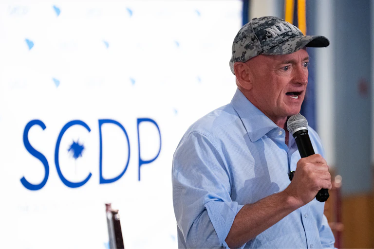 A man in a blue shirt and Navy cap speaks into a microphone.