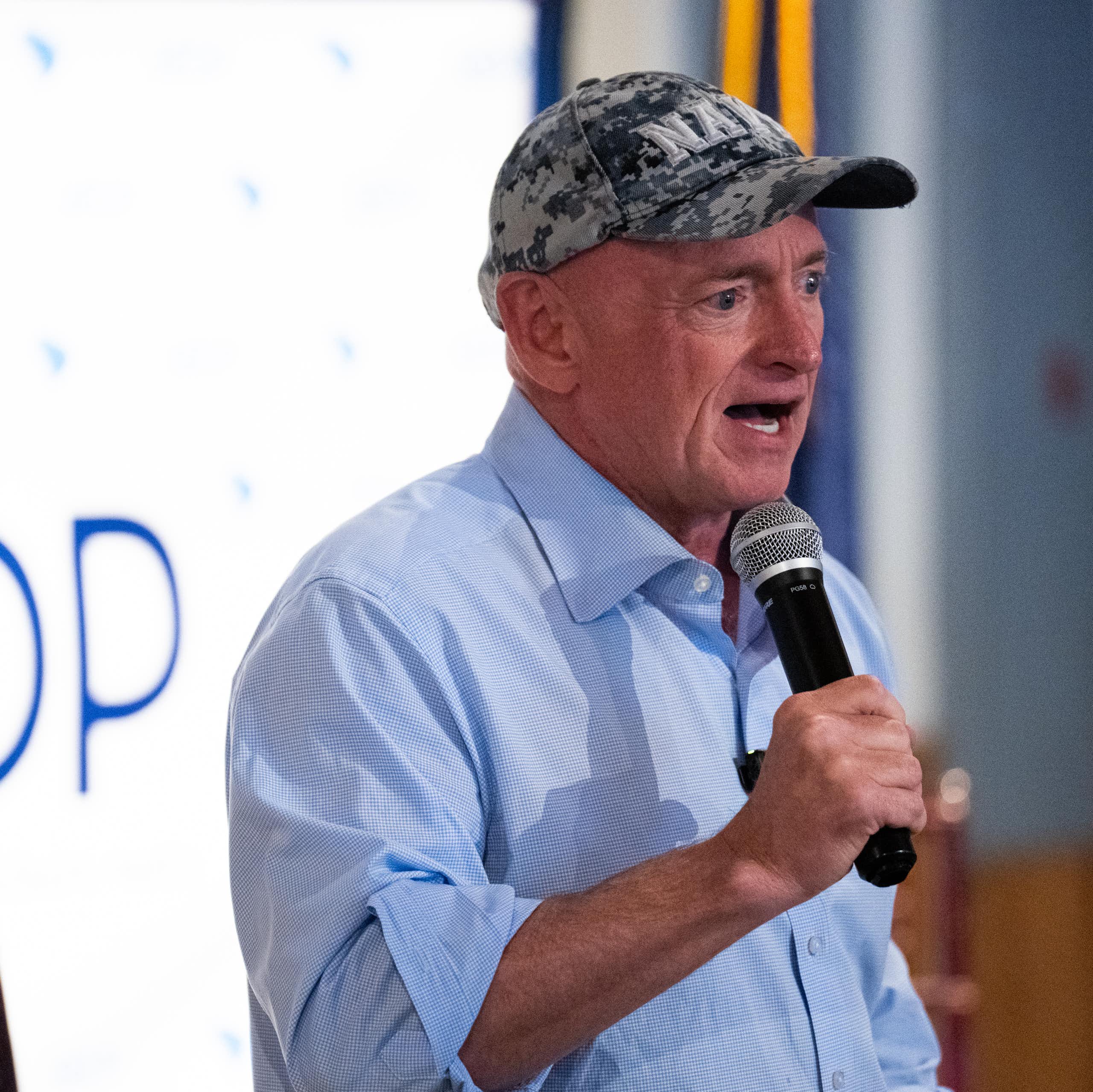 A man in a blue shirt and Navy cap speaks into a microphone.