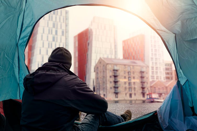 View from inside of a homeless tent. Man in dark dirty cloths sitting by the entrance looking at rich high value district houses.