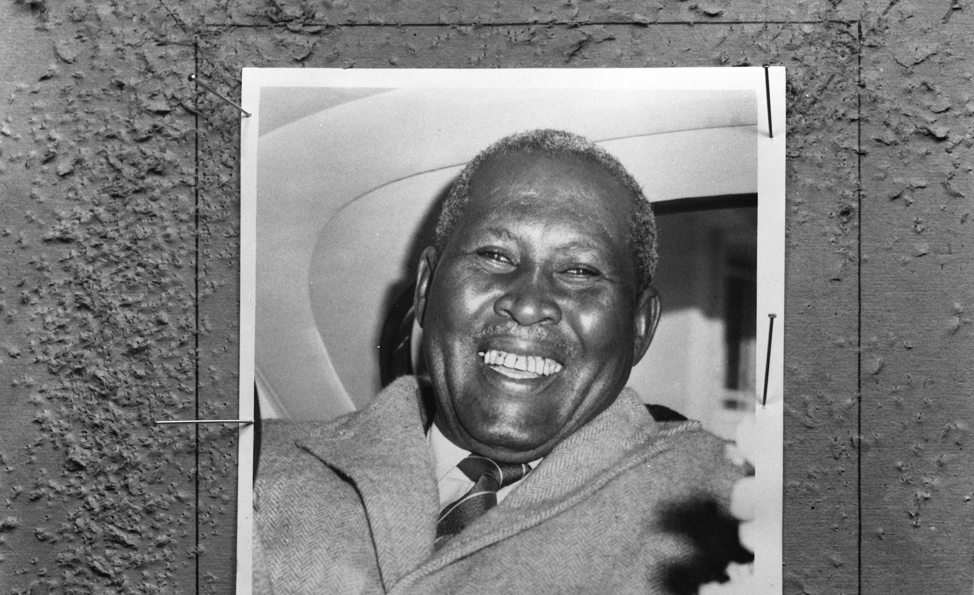 A black and white photo of a smiling African man in the back of a car.