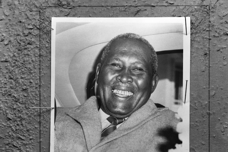 A black and white photo of a smiling African man in the back of a car.