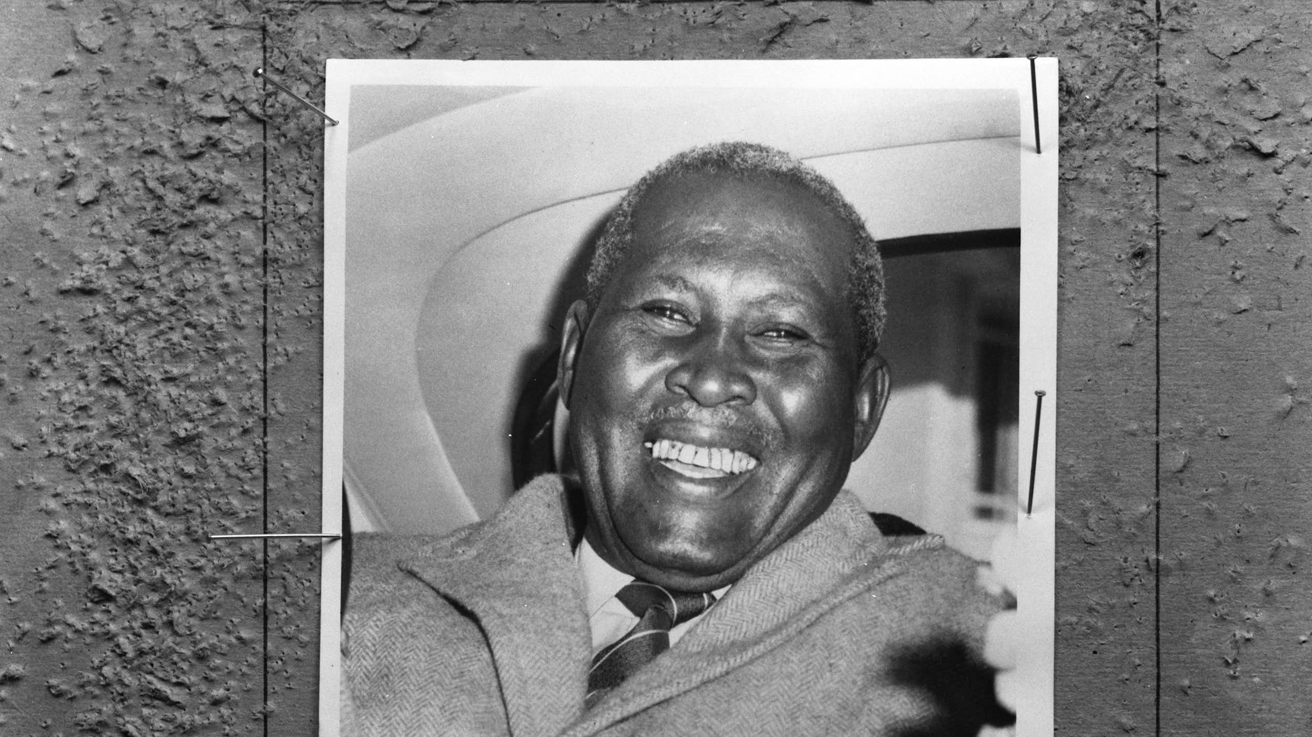 A black and white photo of a smiling African man in the back of a car.