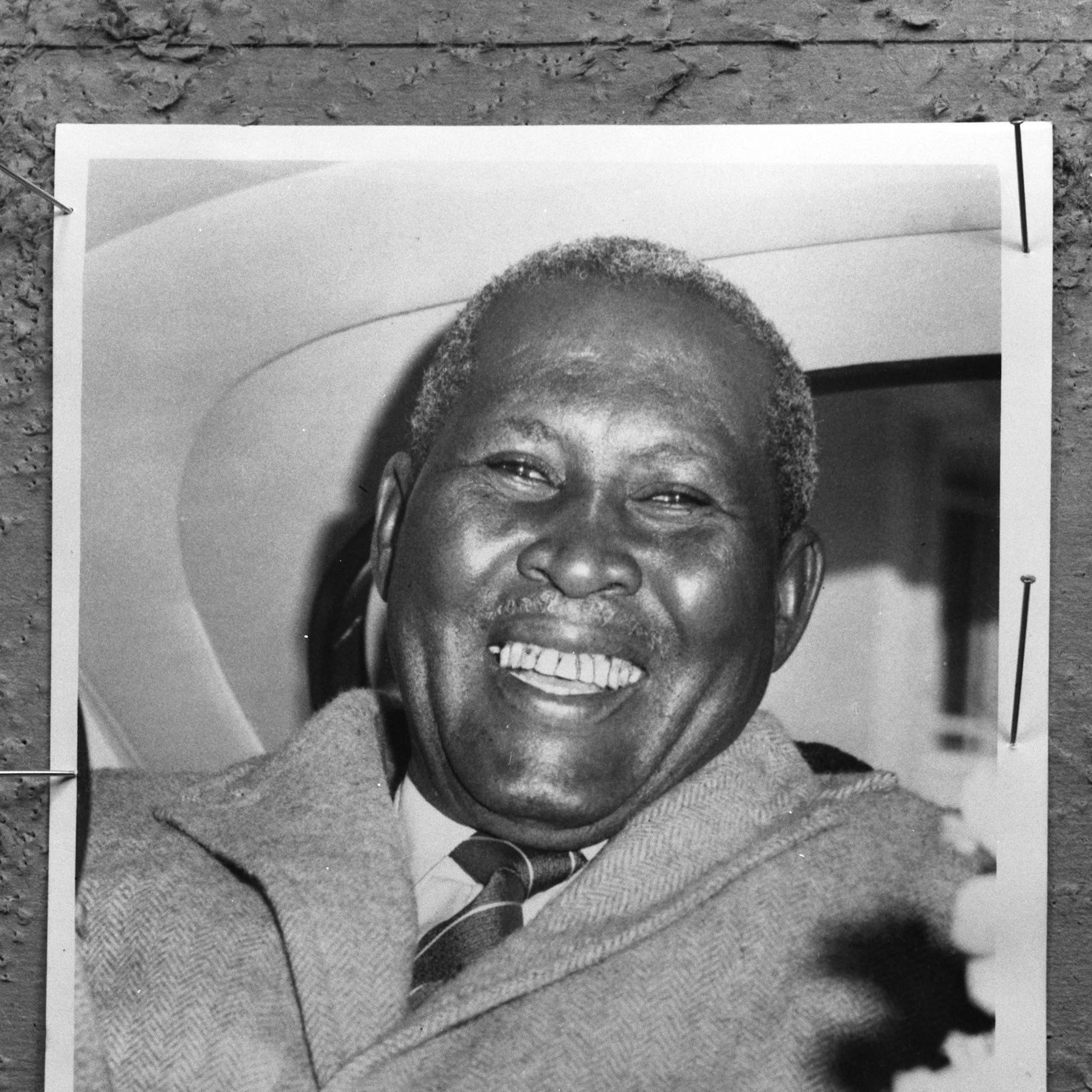 A black and white photo of a smiling African man in the back of a car.