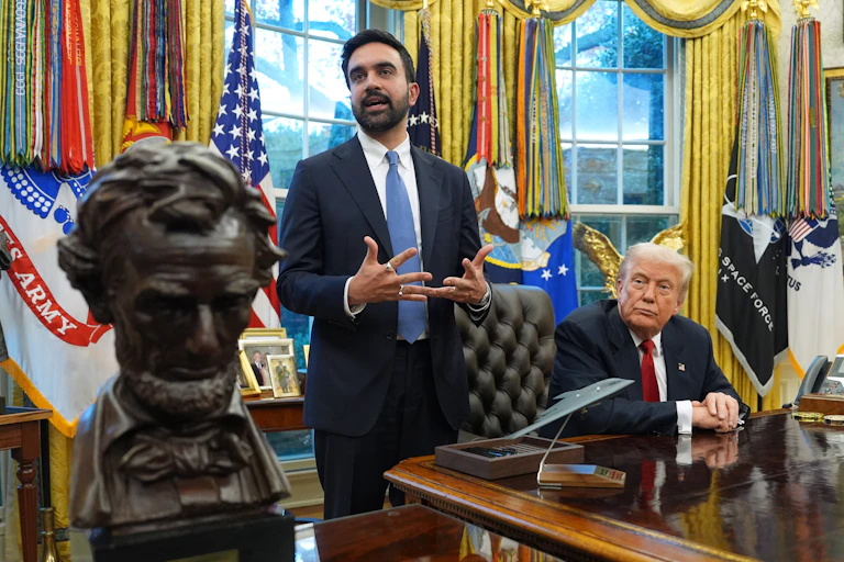 A wooden bust on a desk in front of one standing man in suit and one seated man in suit in front of golden curtains.