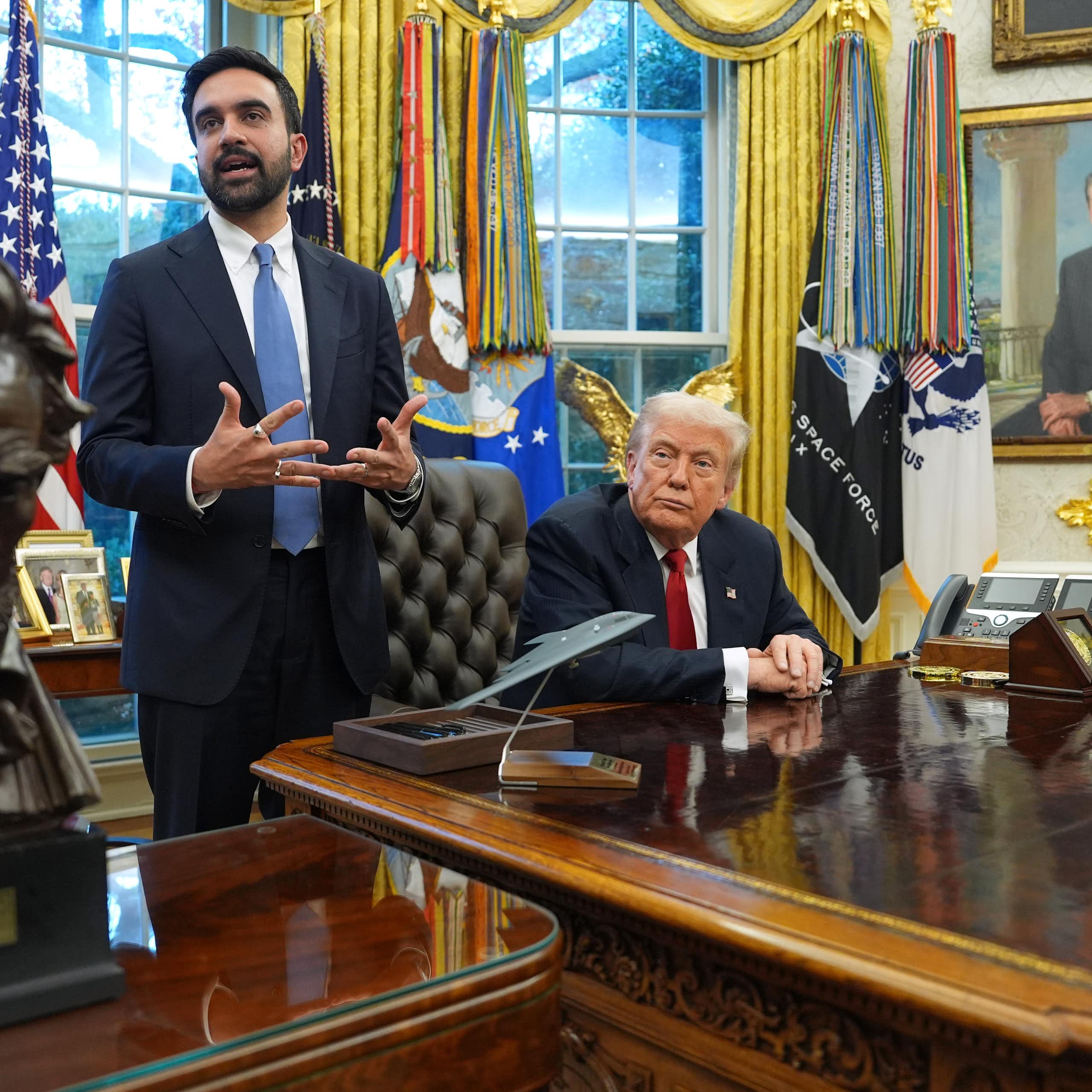 A wooden bust on a desk in front of one standing man in suit and one seated man in suit in front of golden curtains.