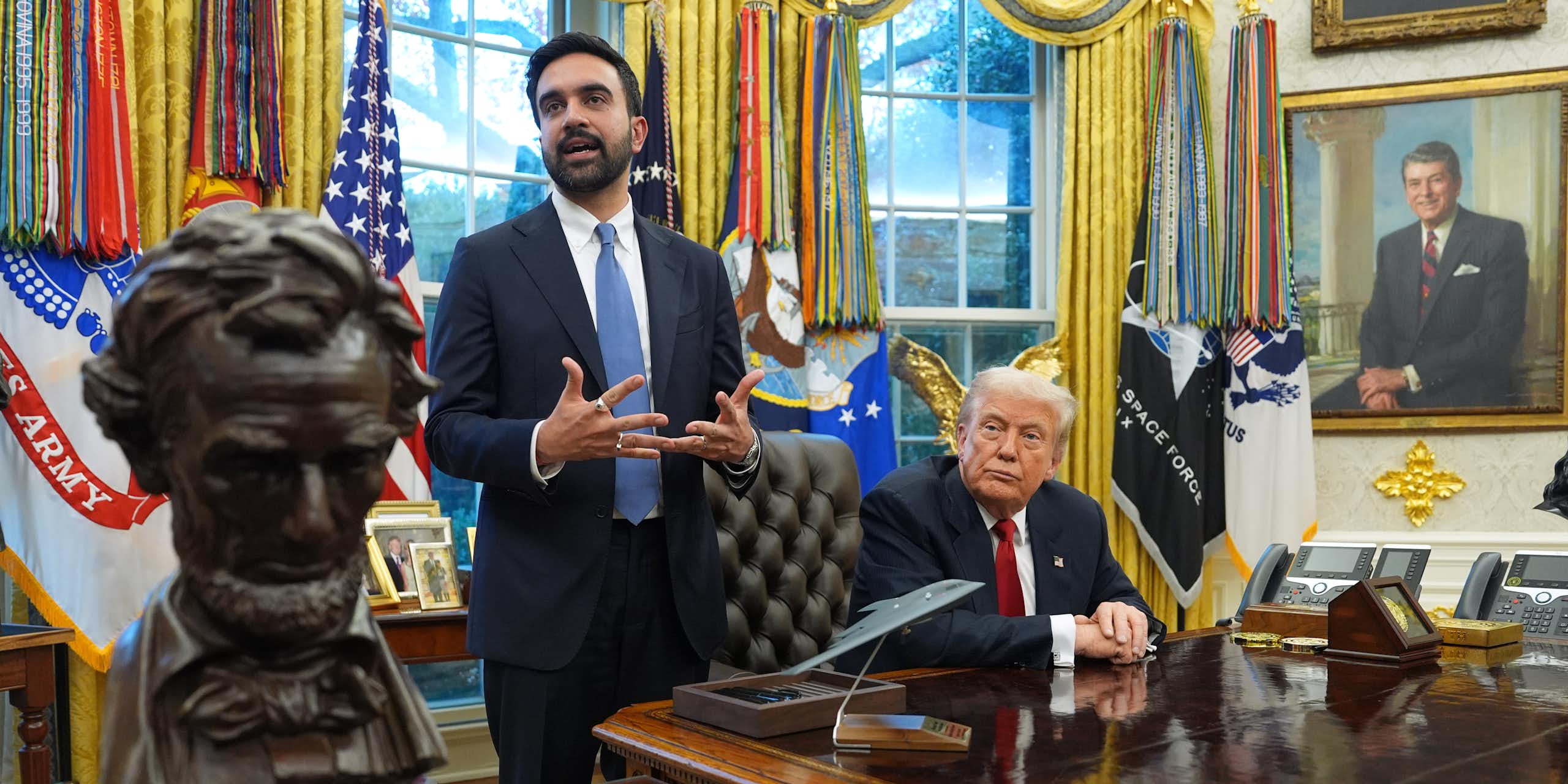 A wooden bust on a desk in front of one standing man in suit and one seated man in suit in front of golden curtains.