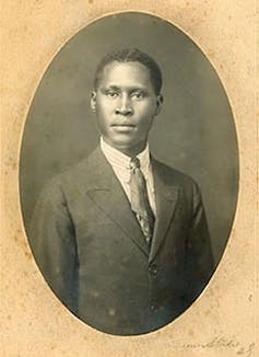 An oval-framed vintage studio photo of a young African man in suit and tie, a middle path in his hair and a proud expression on his face/.
