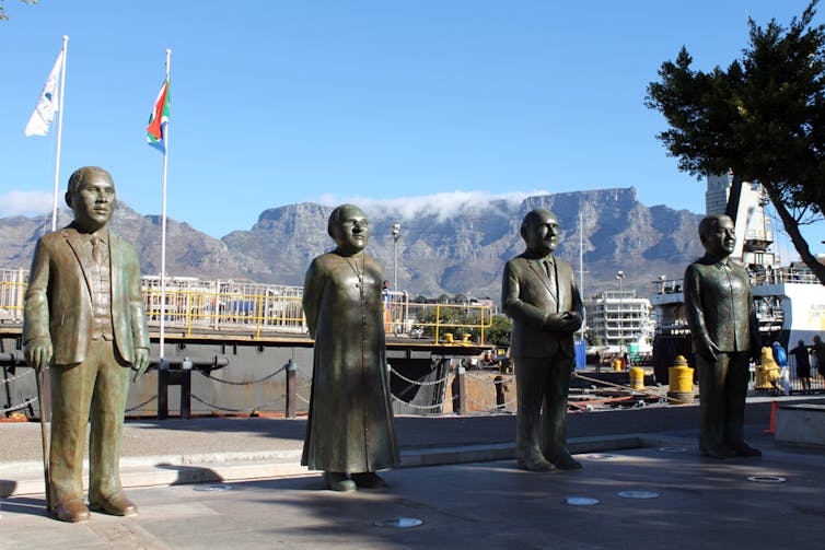 Four statues stand against a backdrop of Table Mountain in Cape Town. They are slightly stylised.