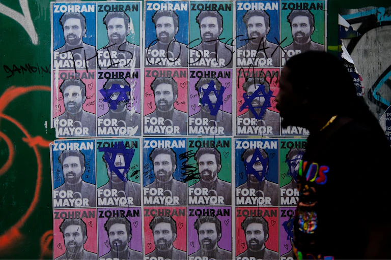 Silhouette of a man walking buy a collage of posters featuring the face of a young, bearded man and the text 'Zohran for Mayor.'