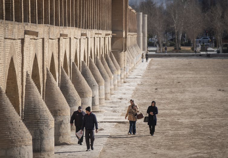 Four people walk next to a bridge across dry ground where a river normally runs.