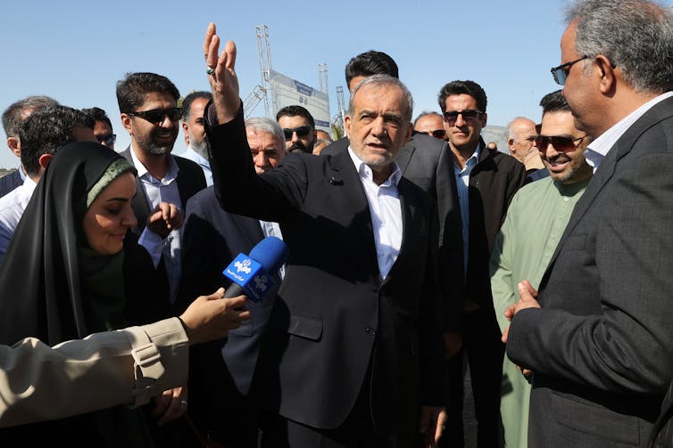 A man gestures while surrounded by other people and speaks into a microphone.