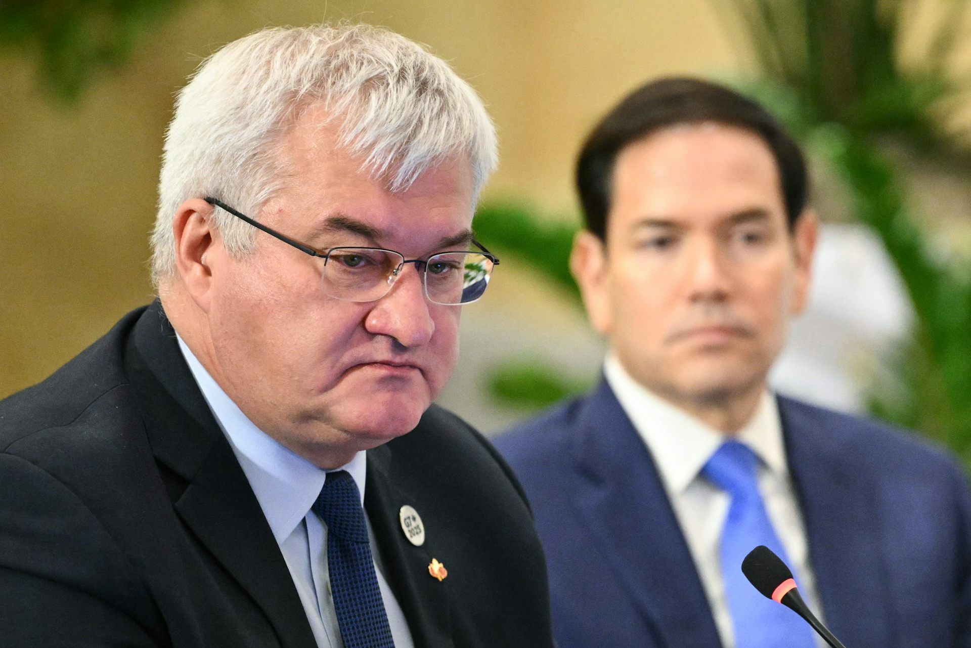 A round-faced grey-haired man wearing glasses with a younger dark-haired man in the background.