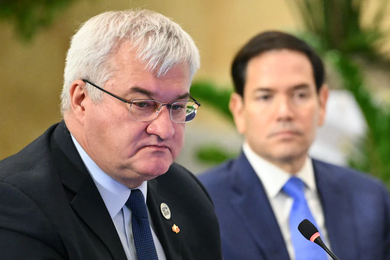 A round-faced grey-haired man wearing glasses with a younger dark-haired man in the background.